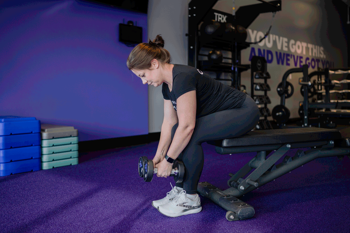 An Anytime Fitness Coach sitting on a bench and demonstrating a seated reverse lateral raise.