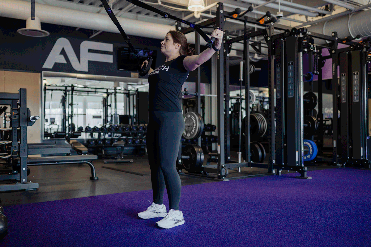 An Anytime Fitness Coach in a large gym performing a TRX reverse fly.