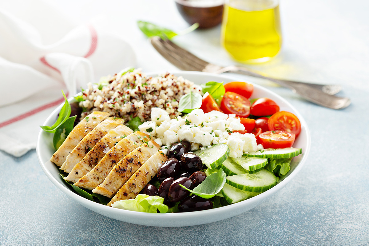 Salad in a bowl, containing chicken, mixed greens, tomatoes, feta, cucumber, olives, and quinoa.