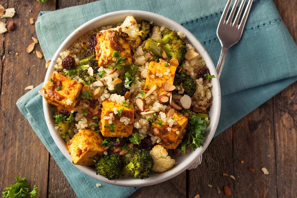 A bowl of quinoa, tofu, and roasted broccoli and cauliflower set on a blue napkin with a fork next to the bowl.